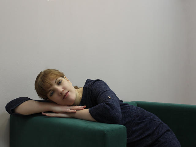 AlissaAmazing is laying on a green couch, wearing a navy blue dress, with her head resting on her arm, looking at the camera with a slight smile.