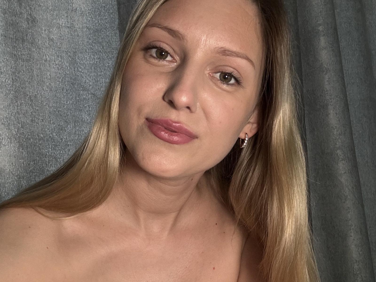 EvaUeslym heeft blond haar, een lichte huid, lichtbruine ogen en roze lippen. Ze glimlacht zachtjes, draagt kleine oorbellen, en haar schouder is zichtbaar. Haar gezicht is gecentreerd..