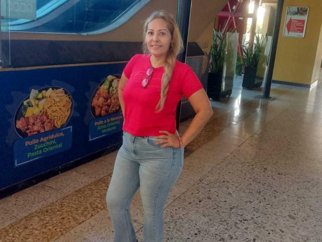 AgataRamos met blonde vlechten, een rood shirt, blauwe jeans, handen op haar heupen, lachend, staand in een gang, met roze zonnebril op haar borst, ziet er schattig uit met haar licht opgetrokken achterwerk..