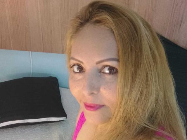 AgataRamos heeft blond haar, bruine ogen en roze lippenstift. Ze draagt een roze top en kijkt naar de zijkant met een lichte glimlach. Haar borsten zijn gedeeltelijk zichtbaar..
