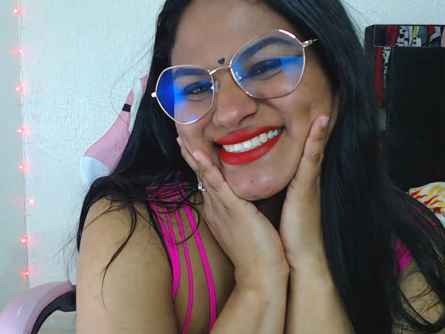 SweetAlma69 with red lipstick, blue-eyed reflection glasses, black hair, pink strappy top, hands on cheeks, smiling, wearing a ring, pink chair in background, fairy lights on wall.