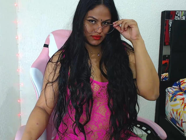 SweetAlma69 with long black hair, pink lace top, red lipstick, and glasses, adjusting glasses while sitting in a pink chair, showing her curvy body and large tits.