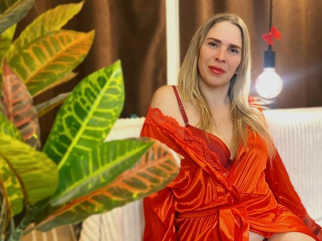 Tessa, con i capelli biondi, rossetto rosso e lingerie di pizzo rossa, seduta su un divano bianco, indossa un abito arancione con una spalla scoperta, con grandi foglie di piante verdi e rosse in primo piano..