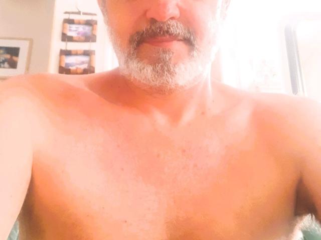 Shirtless DavisBeautiful with a grey beard and mustache, slightly sunburned chest, and a slight smile in a bright room. His nipples are partially visible.