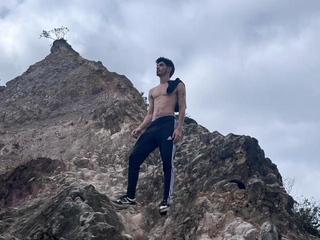 FabrizzioHot, shirtless, with a black backpack, wearing black track pants and black sneakers, standing on rocky terrain, looking to the side with a slight smile, cloudy sky.
