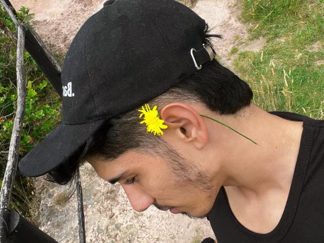 FabrizzioHot with short black hair, black cap, and yellow dandelion in ear, black shirt, gravel ground, green grass, and tree in background. Side profile, looking downward.