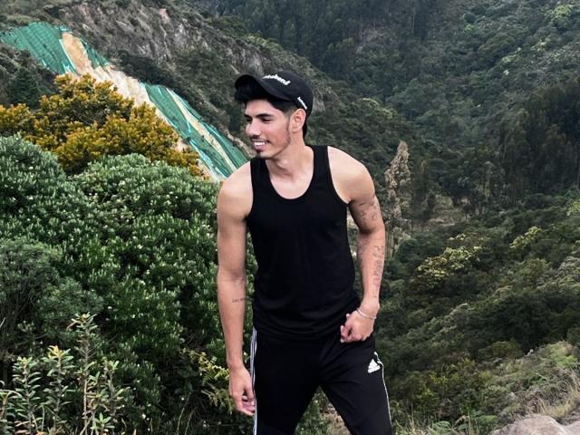 FabrizzioHot, muscular dude with tattoos, black tank top, black pants, black cap, smiling, standing in green forest, looking to the side, hands in pockets, mountain background.