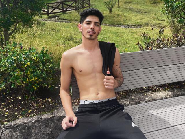 FabrizzioHot, shirtless, with a black sleeveless shirt draped over his shoulder, black Calvin Klein pants, sitting on a wooden bench, tattoos on his right arm, looking at camera.