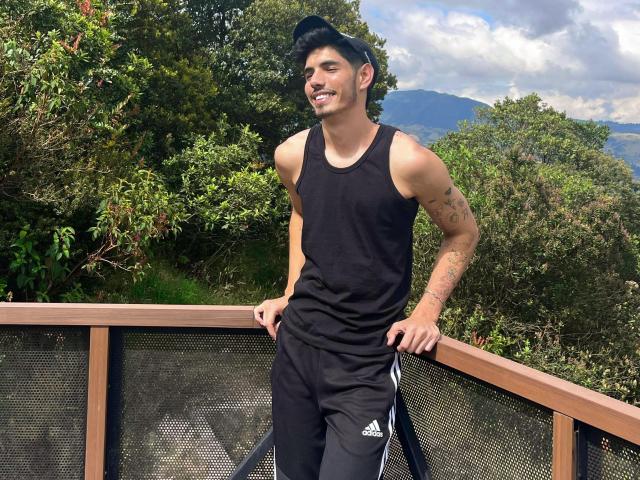 FabrizzioHot, muscular dude with tattoos, black tank top, black cap, black Adidas pants, standing on wooden balcony, hands in pockets, smiling, green forest background, mountains and cloudy sky.