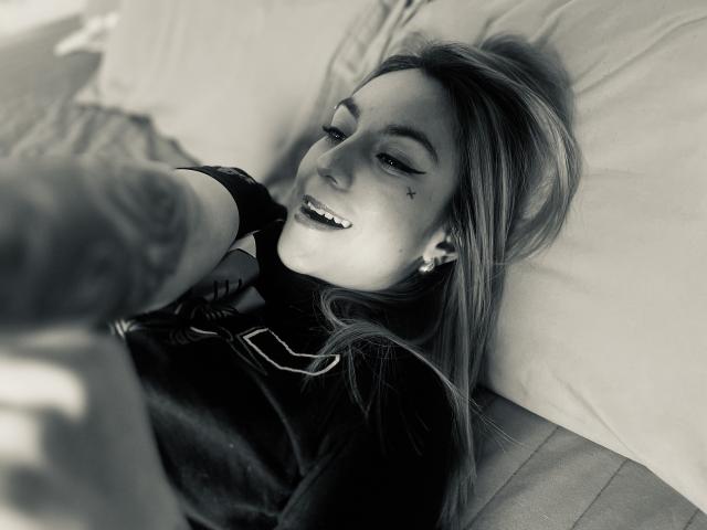 AmyStreetwise69 lying on a pillow, smiling with eyes half-closed, tattoos on her arm, wearing a dark shirt, light hair, small nose, and ear studs, in a black-and-white photo.