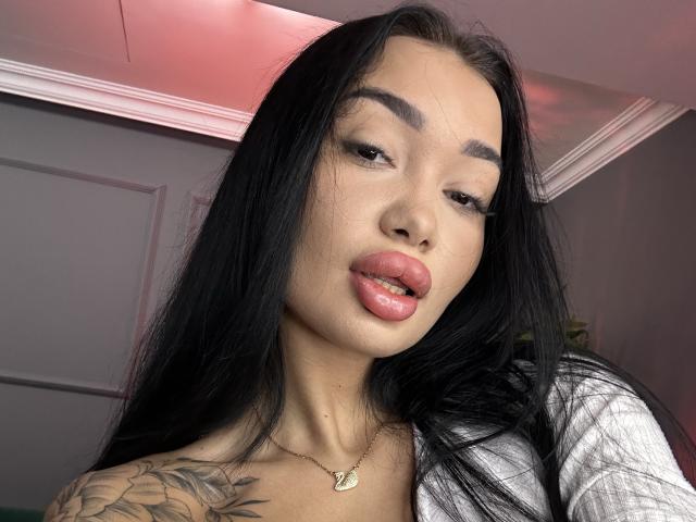Sasha Ferrera tiene el pelo largo y negro, labios rosados y un tatuaje en el hombro derecho. Lleva una blusa blanca y un collar dorado con un pequeño colgante..
