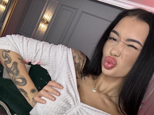 SashaFerrera with black hair, white off-shoulder top, and tattoos on her arm, sticking out her tongue and winking, wearing a gold necklace, showing her tits.