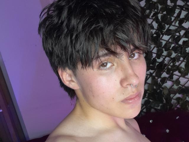 PepperTwink with short black hair, brown eyes, nose ring, and light skin, looking at the camera with a neutral expression, shirtless, in a purple-lit room.