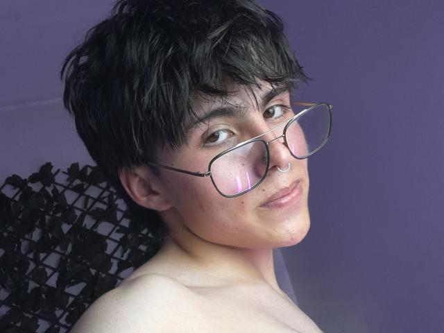 PepperTwink with short black hair, glasses, nose ring, and a slight smile, showing bare shoulders against a purple background. Boobs not visible, but face and upper chest are.