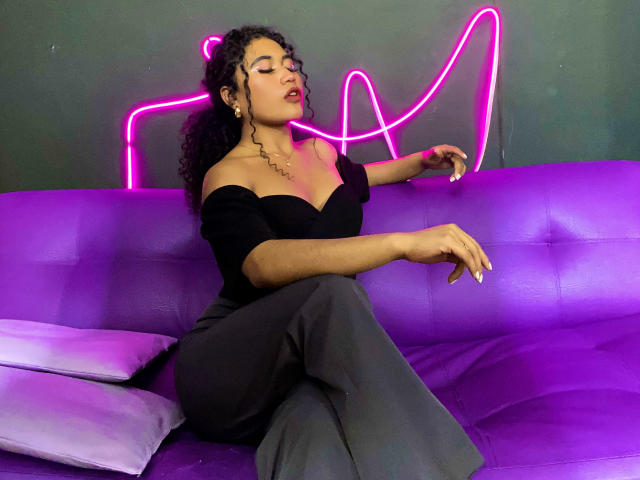 SophieSonyas, avec ses cheveux noirs bouclés, son haut noir à épaules dénudées et son pantalon gris, assise sur un canapé violet, avec un contour néon rose en forme de poisson sur un mur vert foncé, les yeux fermés..