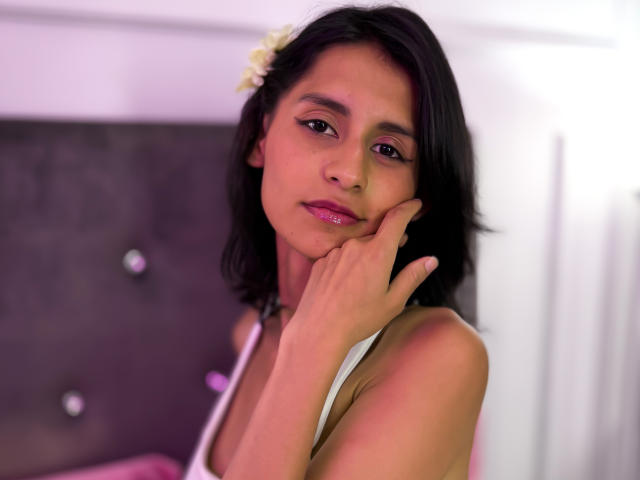 JasminGarcia with short black hair, white flower in hair, pink lipstick, hand on cheek, wearing white tank top, looking at camera, soft pink and white lighting.