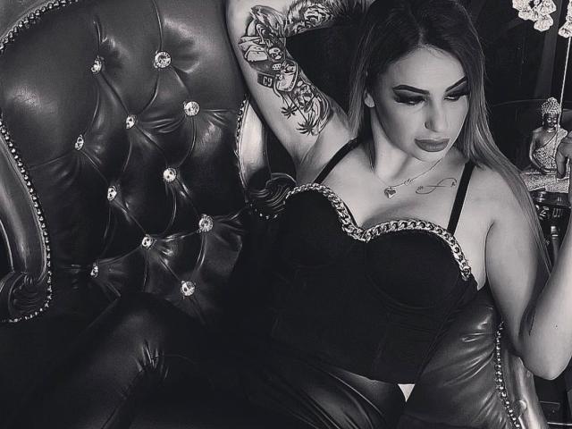 LexiWilld, met tatoeages, een zwarte corset en leren broek, zittend op een gestoffeerde leren stoel, arm omhoog, naar beneden kijkend, waarbij haar borsten en billen zichtbaar zijn, op een zwart-wit foto..