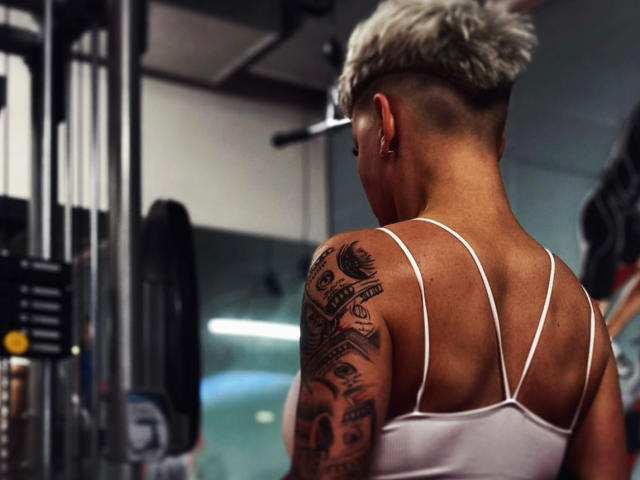 LexiWilld, com os lados da cabeça raspados, um moicano loiro platinado, uma camiseta branca, mostrando os braços tatuados e um corpo musculoso, está em uma academia com aparelhos de musculação..
