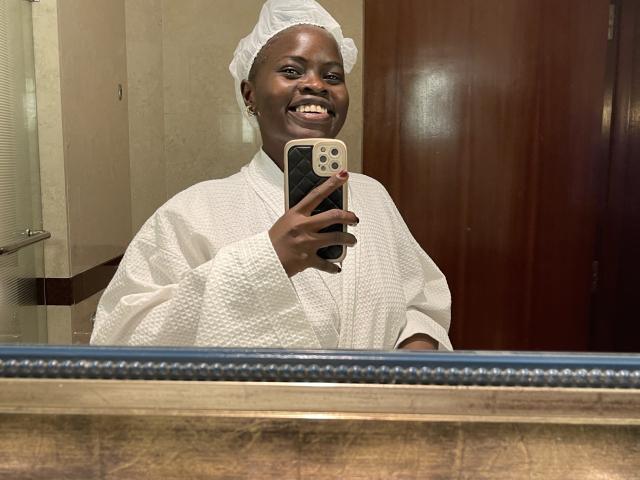 BaddestLinaX in a white towel, smiling with dark skin, wearing a white cap, holding a phone, reflecting in a mirror, showing her teeth and looking happy.