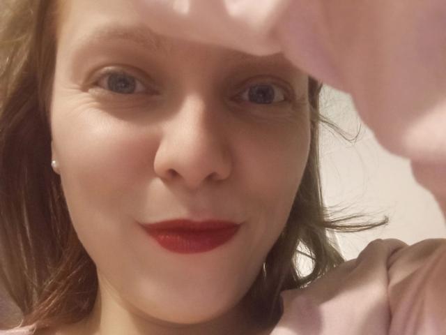JaninaHOT-hot has red lips, blue eyes, and light brown hair, wearing a pink shirt, with a pink shirt sleeve in the foreground. She's smiling subtly.