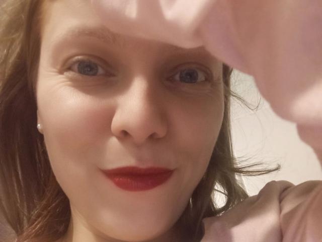 JaninaHOT-hot's face, with red lipstick, blue eyes, and fair skin, wearing a pink shirt, holding her shirt up, showing her nose and smile, with brown hair.