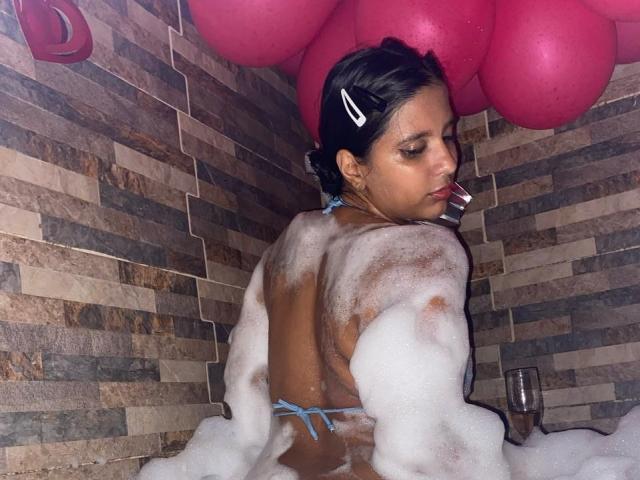 MariaSmith dans une baignoire remplie de bulles, dos à la caméra, avec de la mousse de savon sur son dos et ses fesses nus, cheveux noirs avec une pince blanche, suçant une paille..