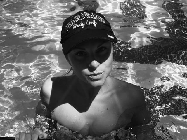 BelleLisaG, in a pool, wearing a baseball cap, with water up to her tits, looking up, sunlight reflecting off the water, black and white photo.