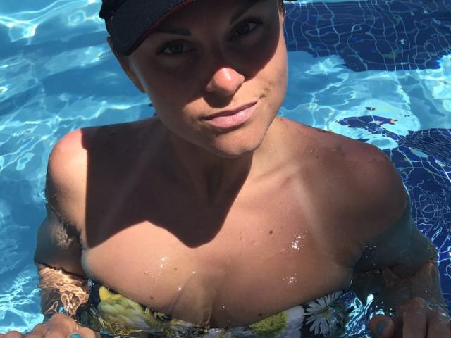 BelleLisaG in a pool, wearing a black cap, with wet, sunlit skin, and yellow floral swimsuit, showing her large, shiny boobs, and tattooed arms, looking up.
