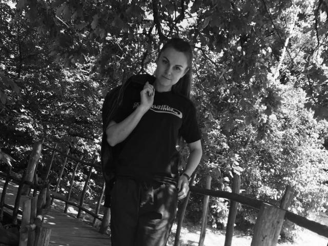 BelleLisaG com cabelo longo e escuro, camiseta preta e calças de couro, segurando uma alça, em pé em uma ponte de madeira, olhando para a câmera, foto em preto e branco..