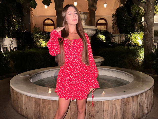 KimberlyWorld, standing in front of a fountain, wearing a red floral dress with long sleeves, her hand touching her brown hair, smiling with her pink lips slightly parted.