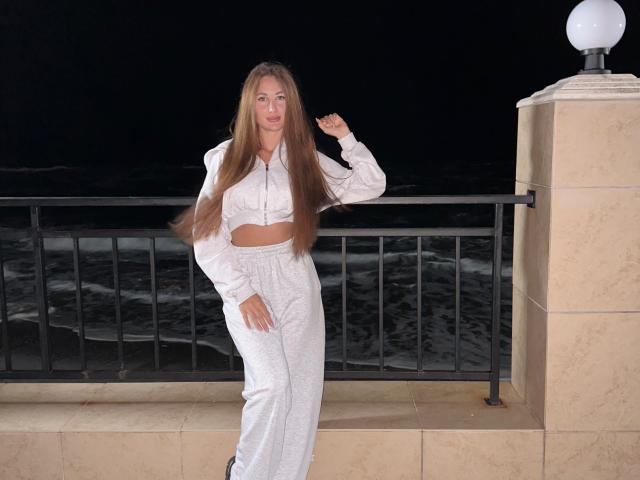 KimberlyWorld with long brown hair, white crop top and matching pants, standing on a balcony at night, hand in hair, waves visible in background, looking relaxed.