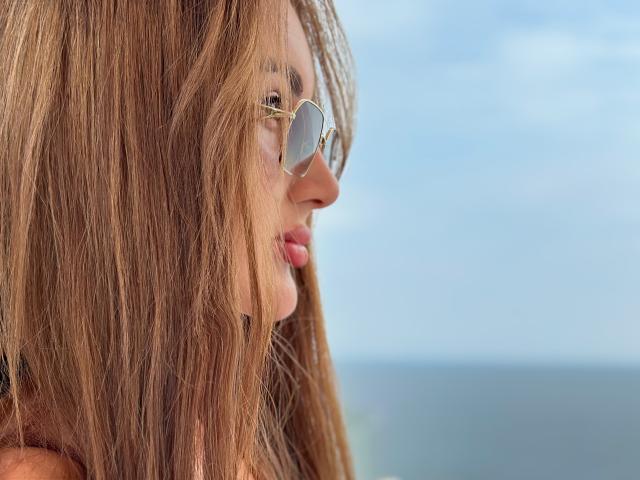 KimberlyWorld with long, straight brown hair, wearing round aviator sunglasses, showing a side profile of her face with pink lips, looking slightly downward.