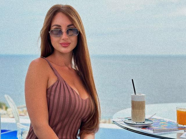 KimberlyWorld, avec de grosses seins, portant un débardeur marron texturé, des lunettes et de longs cheveux bruns, sirotant un café avec une paille, assise à une table avec un magazine..
