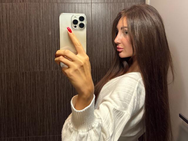 KimberlyWorld, com cabelos castanhos longos, unhas vermelhas, suéter branco texturizado, segurando um iPhone, tirando uma selfie no espelho, mostrando o perfil lateral, com um leve sorriso, fundo de parede de madeira marrom..