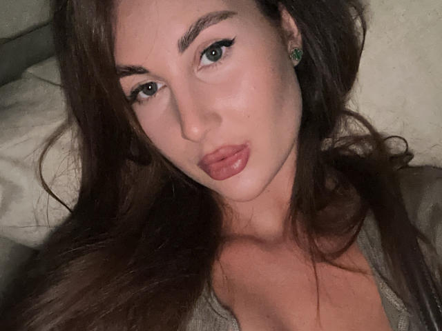 KimberlyWorld a de longs cheveux bruns, des yeux bleus et des lèvres pleines roses. Elle porte des boucles d'oreilles vertes, un haut gris qui dévoile sa poitrine, et est allongée, avec une légère moue sur son visage..