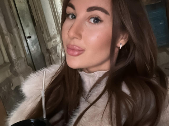 KimberlyWorld has brown hair, blue eyes, and pink lipstick, wearing a white fur coat, with a small diamond earring in her left ear. Her tits are partially visible.