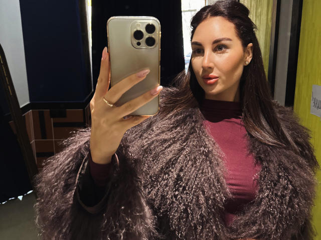 KimberlyWorld mit langem, braunem Haar, rotem Lippenstift und einem flauschigen, violetten Mantel, die ein Selfie mit ihrem iPhone macht und dabei ihre Brüste in einem burgunderfarbenen Rollkragenpullover zur Schau stellt..