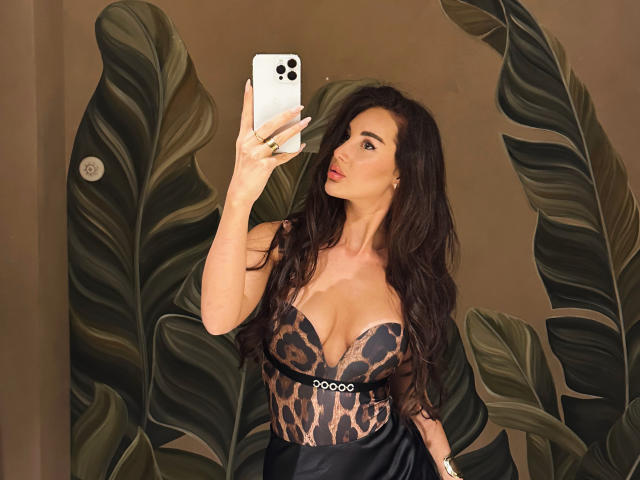KimberlyWorld with long dark hair, wearing a leopard-print top, taking a selfie with her iPhone, showing her big boobs and red lipstick, against a brown wall.