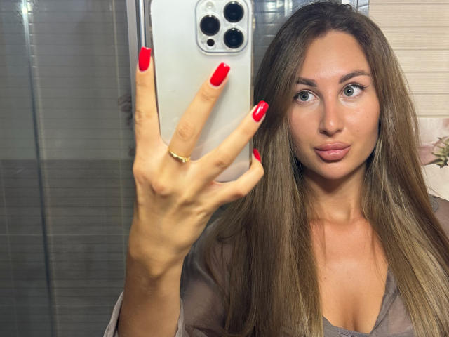 KimberlyWorld, com cabelos castanhos longos, unhas vermelhas e um anel de ouro, tirando uma selfie com um smartphone, mostrando seu rosto e mão, em um espelho. Seios visíveis em uma blusa cinza..