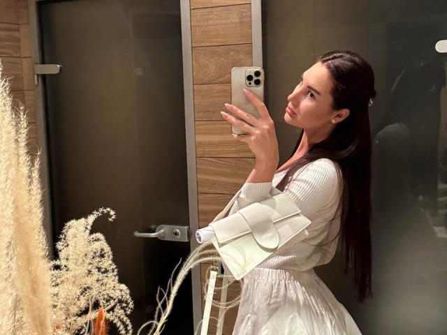 KimberlyWorld mit langem, dunklem Haar, in einem weißen Kleid, hält ein iPhone, macht ein Selfie vor einem Spiegel und zeigt ihre Brüste im Spiegelbild, mit weißen, voluminösen Blumen im Vordergrund..