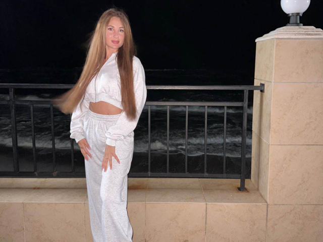 KimberlyWorld met lang, golvend blond haar, gekleed in een witte crop top en bijpassende losse broek, staat 's nachts bij een zwarte reling, met haar hoofd lichtjes gekanteld en een speelse glimlach..