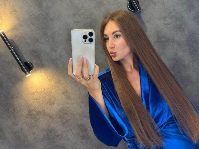 KimberlyWorld, com cabelos castanhos longos, vestindo um robe de veludo azul, segurando um iPhone, com lábios curvados para baixo, um anel no dedo, sob luzes de parede, olhando no espelho, tirando uma selfie. Seios e nádegas não estão visíveis..