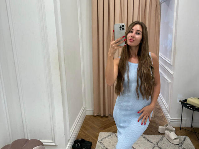 KimberlyWorld in a light blue, sleeveless, form-fitting dress with long brown hair, red nails, and white heels, taking a mirror selfie with a confident smile.