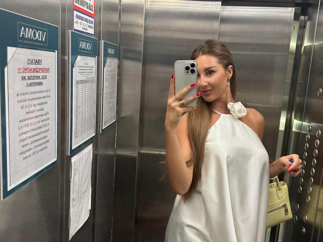 KimberlyWorld mit langem, braunem Haar, weißem Top mit Halter und einem Blumenaccessoire, hält ein Handy in einem silbernen Aufzug, zeigt ihre Brüste und trägt roten Nagellack, hält eine beige Tasche..