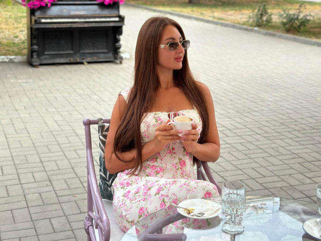 KimberlyWorld met lang bruin haar, gekleed in een bloemenjurk, met een kop in haar hand, zittend in een stoel, met een zonnebril op, kijkend naar de zijkant, met een tafel en een glas voor zich..