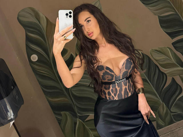 KimberlyWorld, con lunghi capelli scuri, indossa un corpetto con lacci che mette in mostra il seno e una gonna nera, e sta scattando un selfie allo specchio con un iPhone. Al polso ha un orologio..