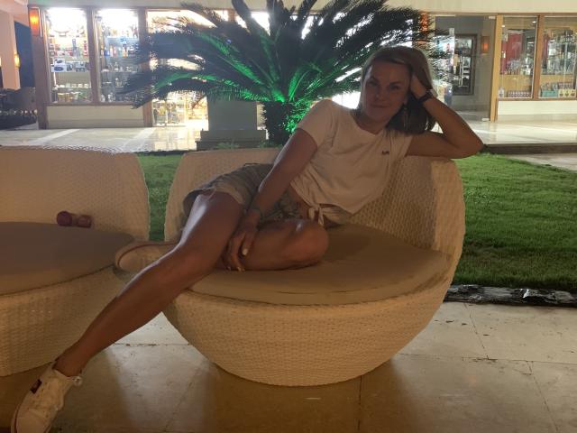 BelleLisaG with tan skin, blonde hair, small tits, white tee, denim shorts, white sneakers, sitting on wicker chair, leg stretched out, hand on head, relaxed, green plant behind.