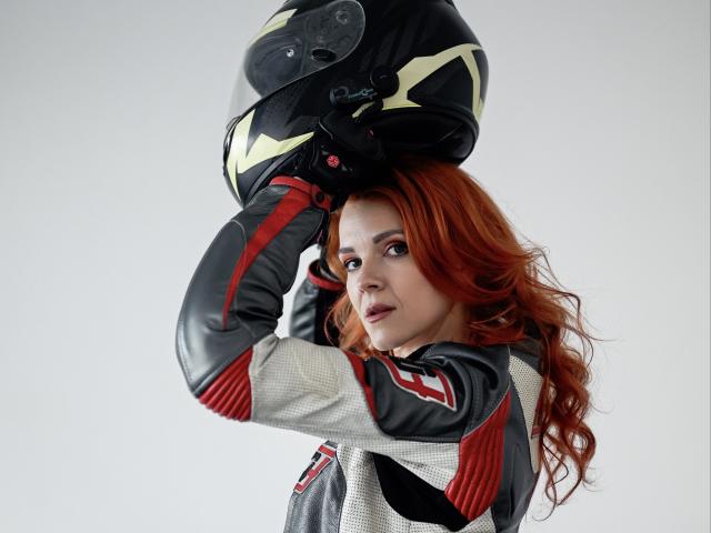 GingerLizzi, com cabelo ruivo, vestindo um macacão de motocicleta cinza e vermelho, colocando um capacete preto com listras amarelas, olhando ligeiramente para o lado com uma expressão concentrada..