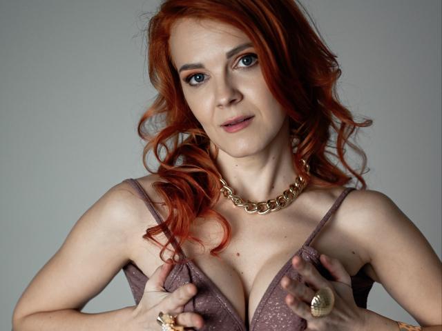 GingerLizzi with red hair, wearing a sparkly purple bra, holding her tits with hands, gold chain necklace, and rings, looking at camera with a slight smile.