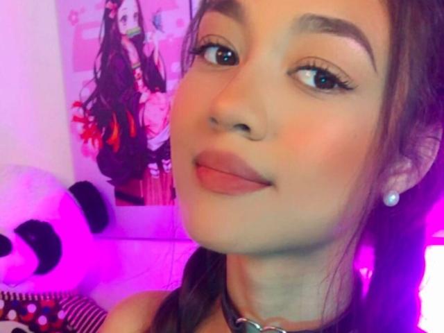 SammyStar69, con labios fruncidos, cejas oscuras y piel rosada, lleva un collar y tiene un piercing en la nariz, con el cabello largo y castaño, luciendo adorable con iluminación rosa y morada..